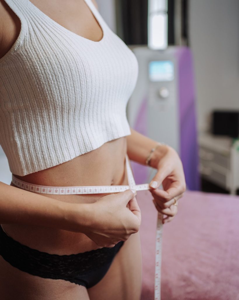 Eine Frau in einem weißen Oberteil misst mit einem Maßband ihren Taillenumfang und unterstreicht damit ihr Engagement für die Gesundheit.