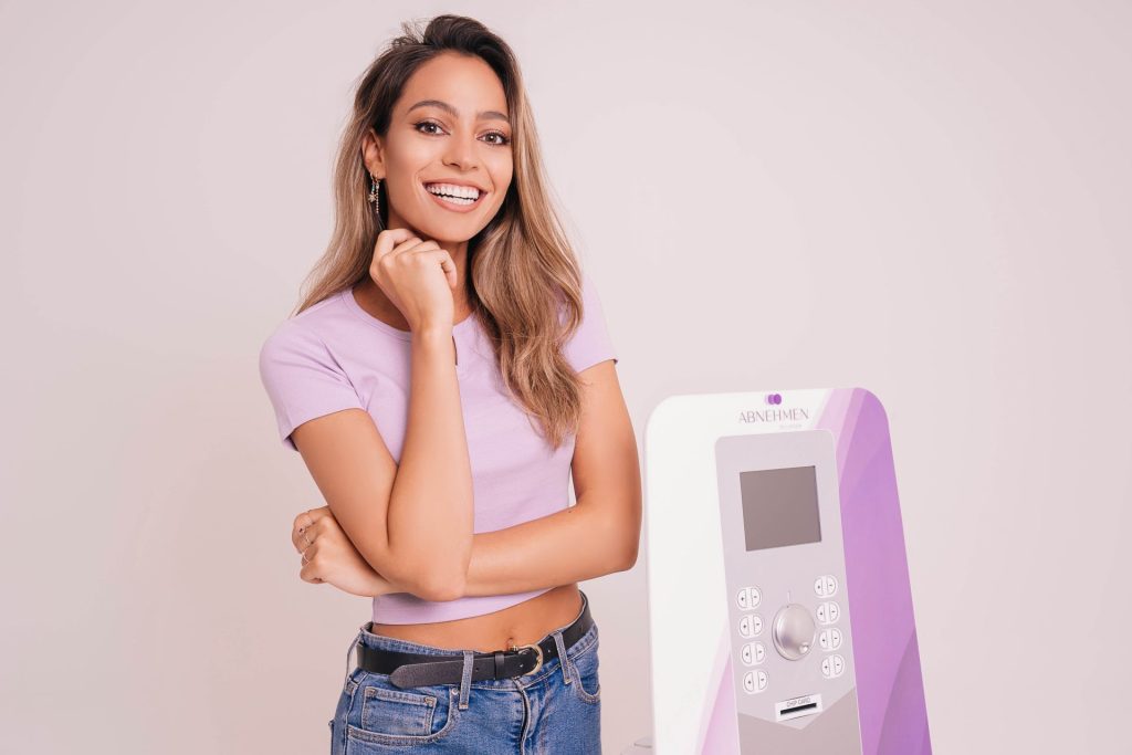 Eine Frau steht neben einem lila-weißen Verkaufsautomaten, der verschiedene Snacks und Getränke anbietet.